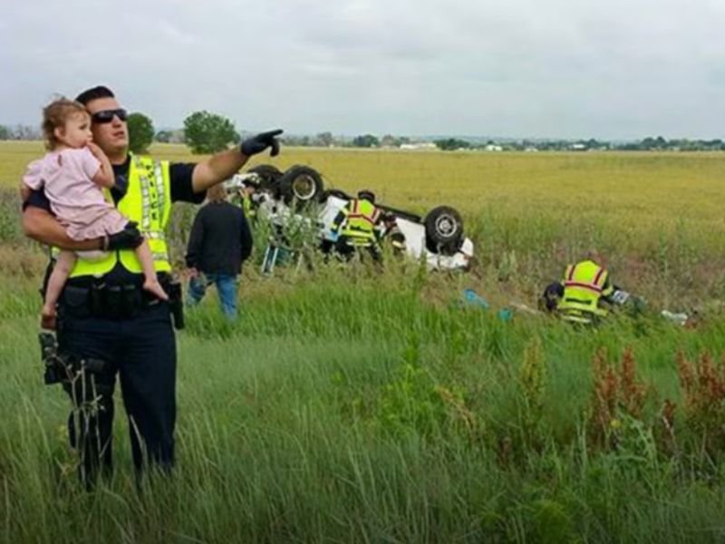 Police Officer Gently Holds A Little Baby Girl, Distracting Her From The Wreckage Where Her Father His Life In A Car Crash
